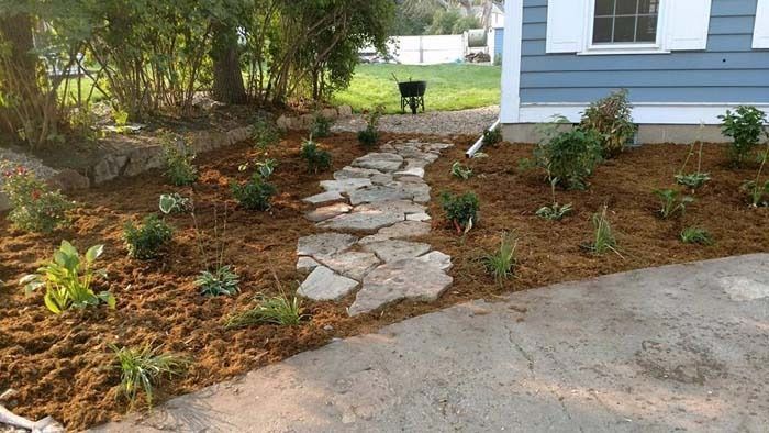 A stone pathway cuts through a mulched garden bed next to a blue-sided house.