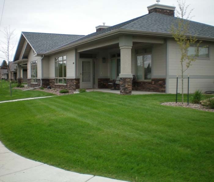 A beige house with stone accents, a covered porch, and a manicured green lawn under an overcast sky.