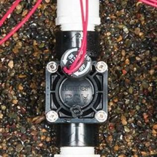 A black Hunter irrigation valve installed in a gravel landscape, featuring a solenoid with red wires and an on/off dial.