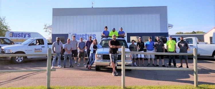 A group of employees stands in front of a Dusty's company building and several service trucks on a sunny day.