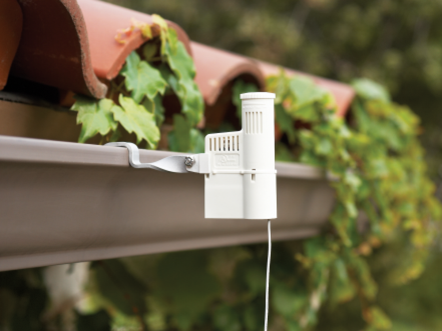 A white rain sensor mounted to the edge of a tan roof gutter, with green foliage and terra cotta roof tiles in the background.