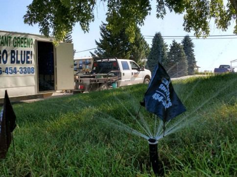 A lawn sprinkler with a branded flag waters grass near a white service truck and a trailer with advertisement text.