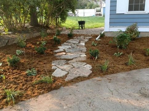 A flagstone path winds through a mulched garden bed next to a blue-sided house with a concrete driveway in the foreground.