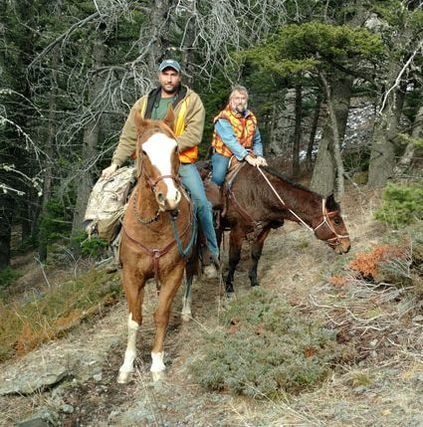 Two people on horseback ride along a dirt trail through a coniferous forest.