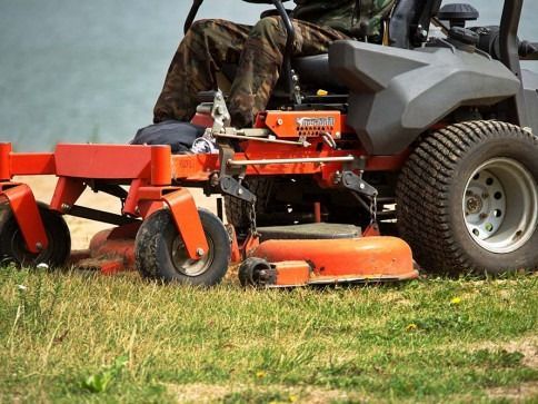 A person operates an orange zero-turn mower on grass near a body of water.