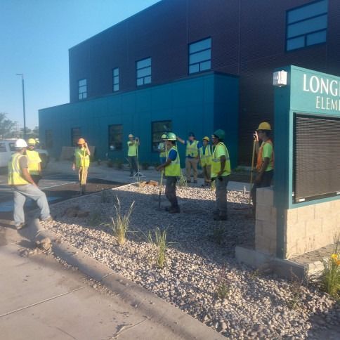 A group of workers in high-visibility vests and hard hats stand outside Longfellow Elementary School on a sunny day.