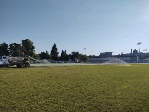 A water truck sprays a wide arc of water across a large green sports field on a sunny day.