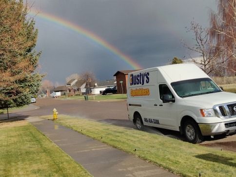 A white delivery van parked on a grassy curb in a suburban neighborhood under a rainbow in a stormy sky.