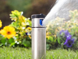 A silver metal lawn sprinkler spraying water over a green yard with yellow and purple flowers in the background.
