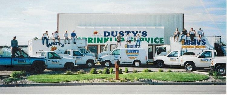 A large group of people sit on the roofs of multiple white service trucks in front of a Dusty's Sprinkler Service building.