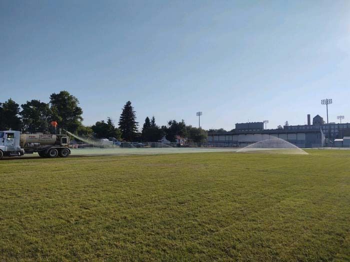 A water truck sprays a fine mist over a large, grassy field on a sunny day.