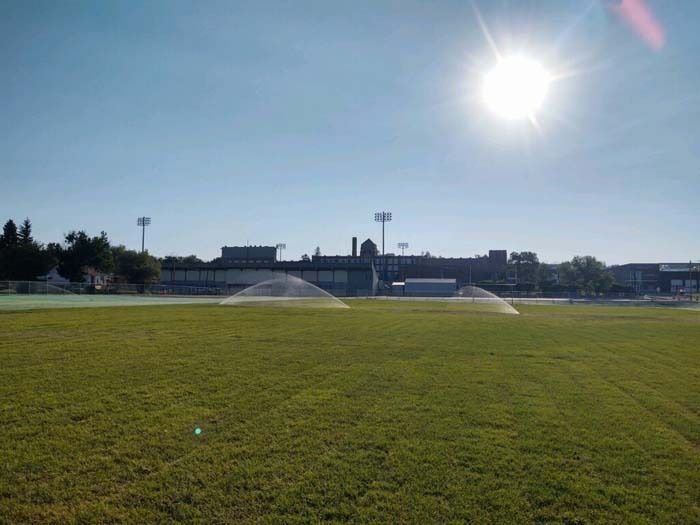 A bright, sunny day over a green field with sprinklers active, featuring a large building and light poles in the distance.