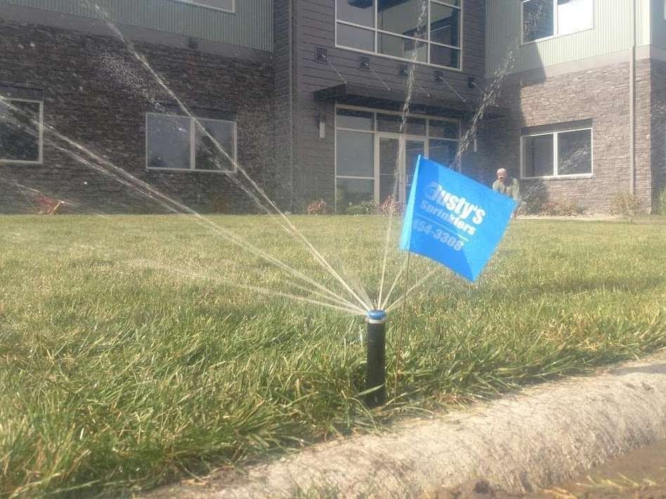 A sprinkler sprays water onto a green lawn in front of a building, marked by a small blue flag for Dusty’s Landscaping.