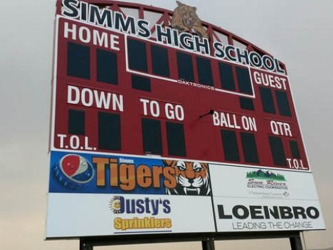 A red and dark-screen Simms High School scoreboard outdoors with advertisements for Tigers, Dusty's Sprinklers, and Loenbro.
