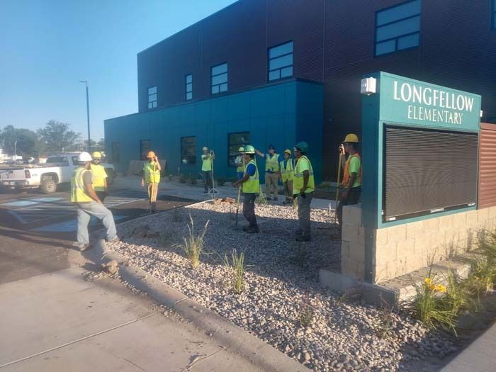 Construction workers in hard hats and safety vests stand outside a Longfellow Elementary School building with blue walls.