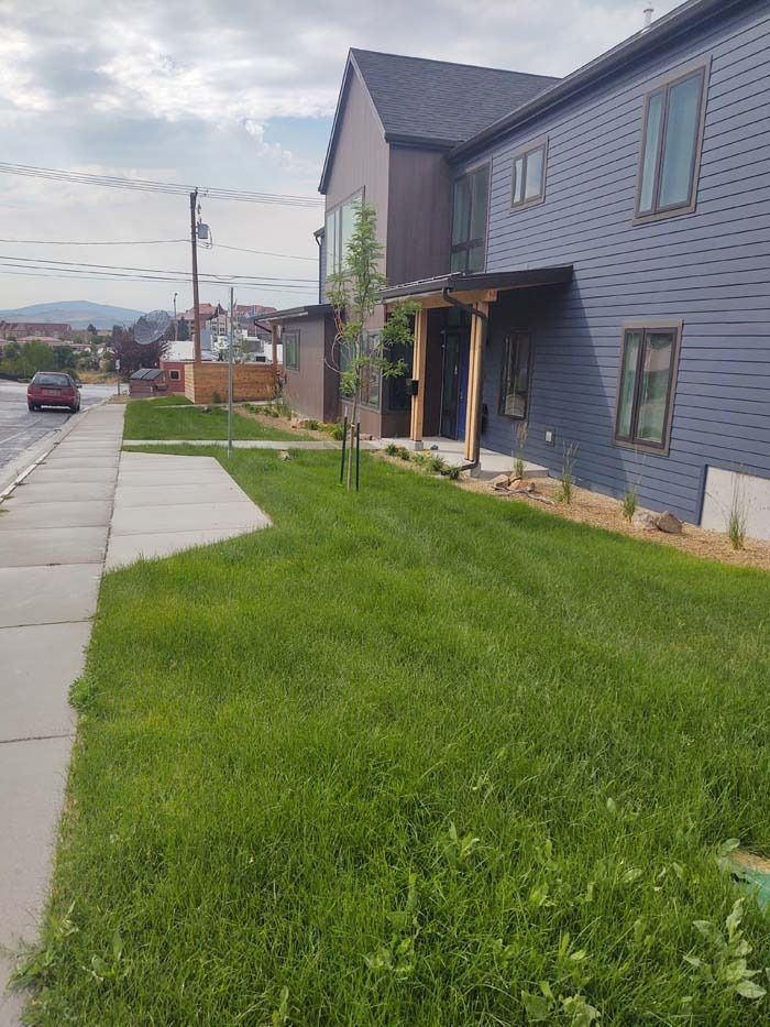 A blue two-story residential building with a wooden porch and a grassy front yard next to a concrete sidewalk.