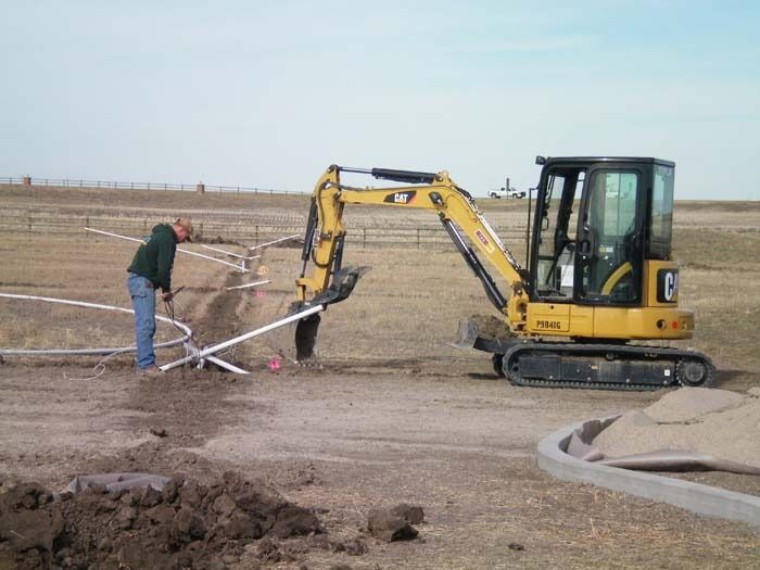A person works on pipes in a trench while a yellow Caterpillar excavator stands nearby on a rural, open field.