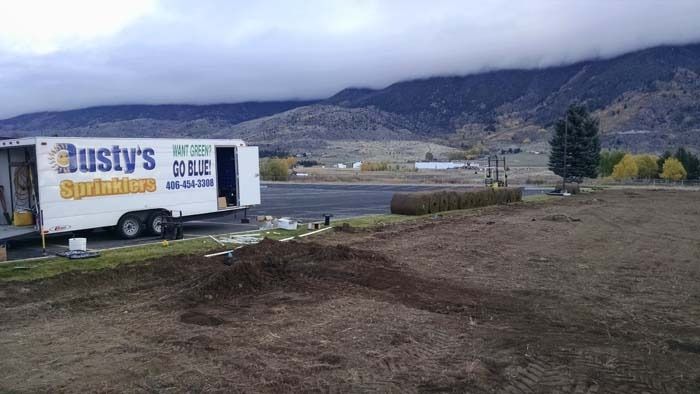 A white Rusty's Sprinklers trailer parked on a dirt lot near a field with mountains in the background under a cloudy sky.