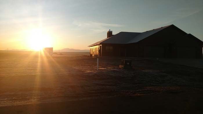 A silhouette of a farmhouse stands in a vast, empty field during a bright sunrise.