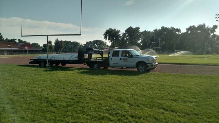 A white truck pulling a long trailer with a white cover, parked on a dirt track by a grassy field with goal posts.
