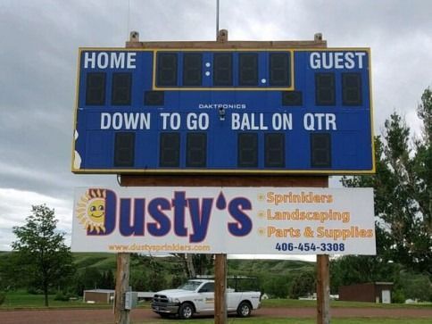 A blank electronic football scoreboard mounted on posts above a Dusty's advertising sign with a white truck parked below.
