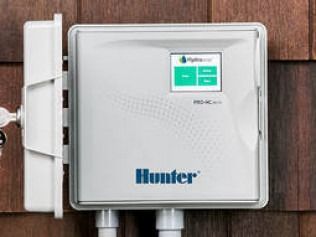 A light gray Hunter Pro-HC outdoor irrigation controller mounted on a brown shingled wall with a key in the side lock.