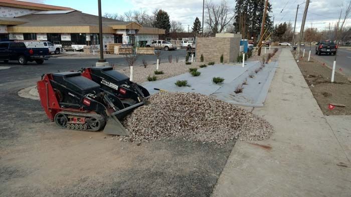 A red track loader pushes a pile of gravel onto a landscaped area next to a building and sidewalk.