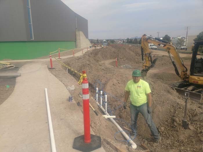 A worker in a neon yellow shirt stands in an excavated construction trench next to a parked yellow excavator.