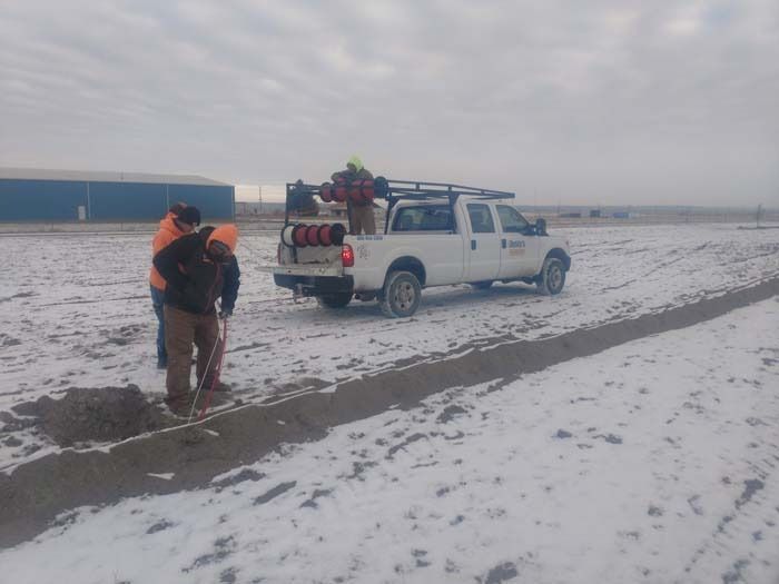 Workers stand by a white service truck in a snowy field, digging in the ground near a trench.