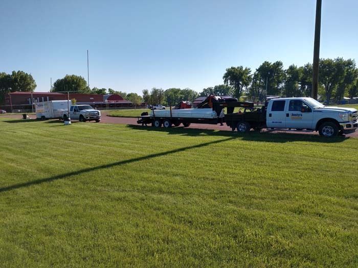 A white truck towing a long trailer with a boat sits on a grass lawn near a parking area with a white trailer.