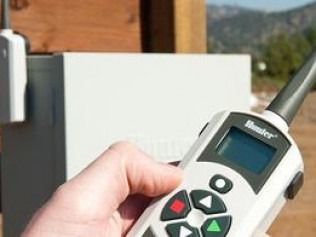 A hand holding a white Hunter Industries remote control in front of an outdoor irrigation controller.