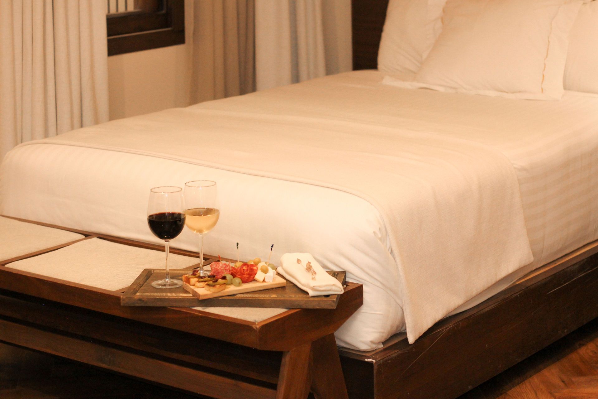 Cama de hotel con sábanas blancas y una bandeja de desayuno con vino y fruta a los pies de la cama.
