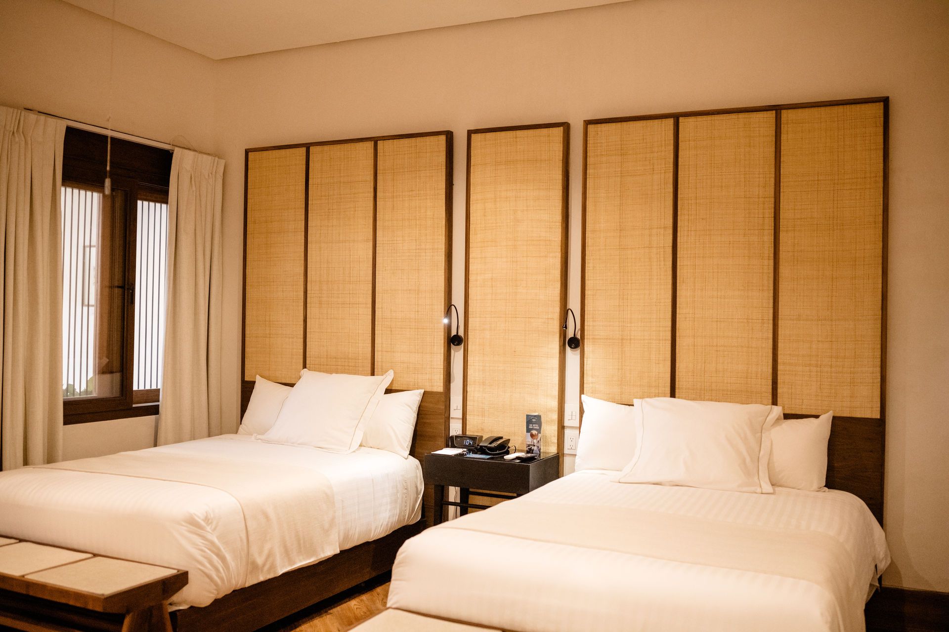 Habitación de hotel con dos camas individuales, cabeceros de bambú y una pequeña mesa compartida entre ellas.