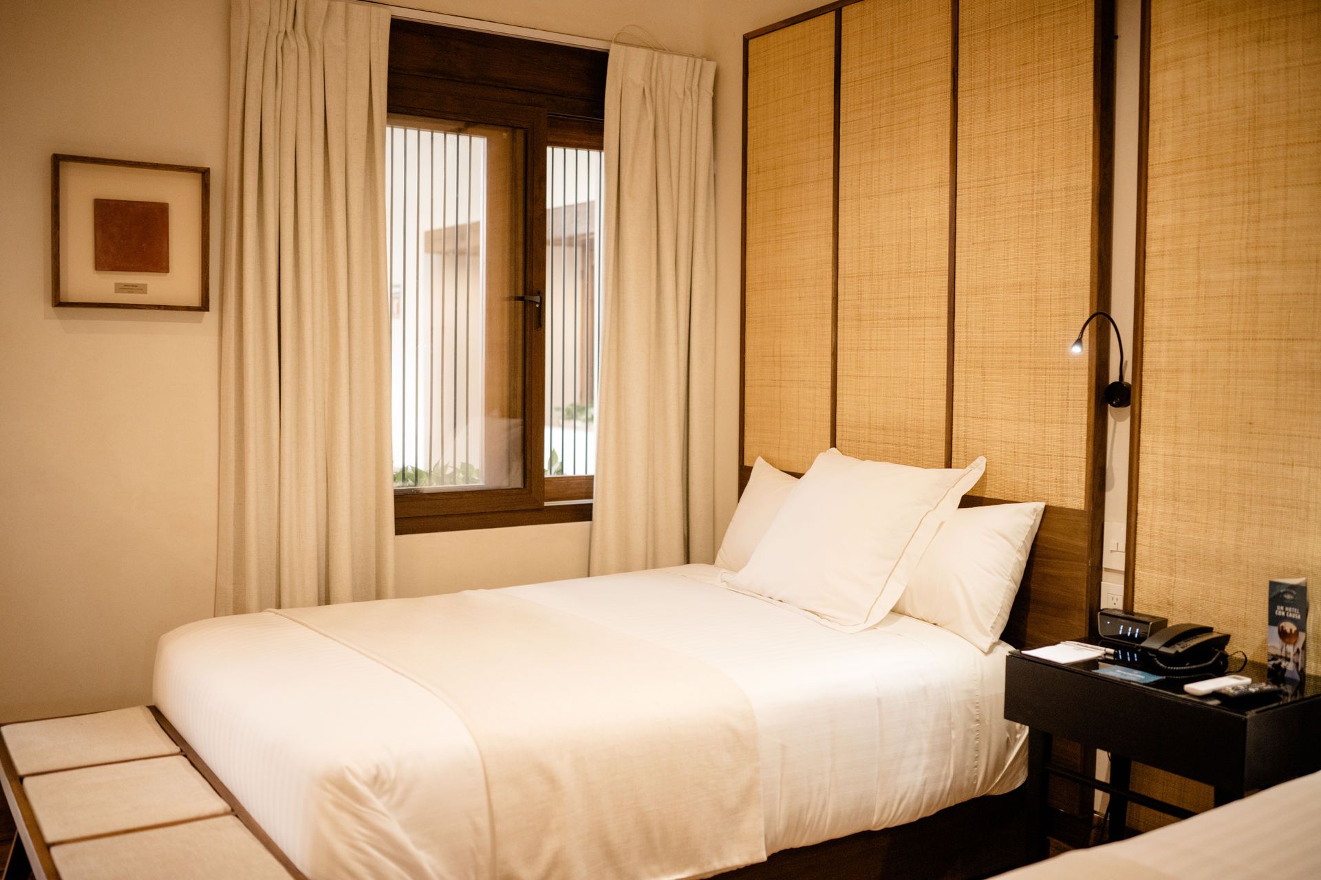 Acogedora habitación de hotel con una cama bien hecha, ventana con cortinas y una mesita de noche con teléfono y lámpara.