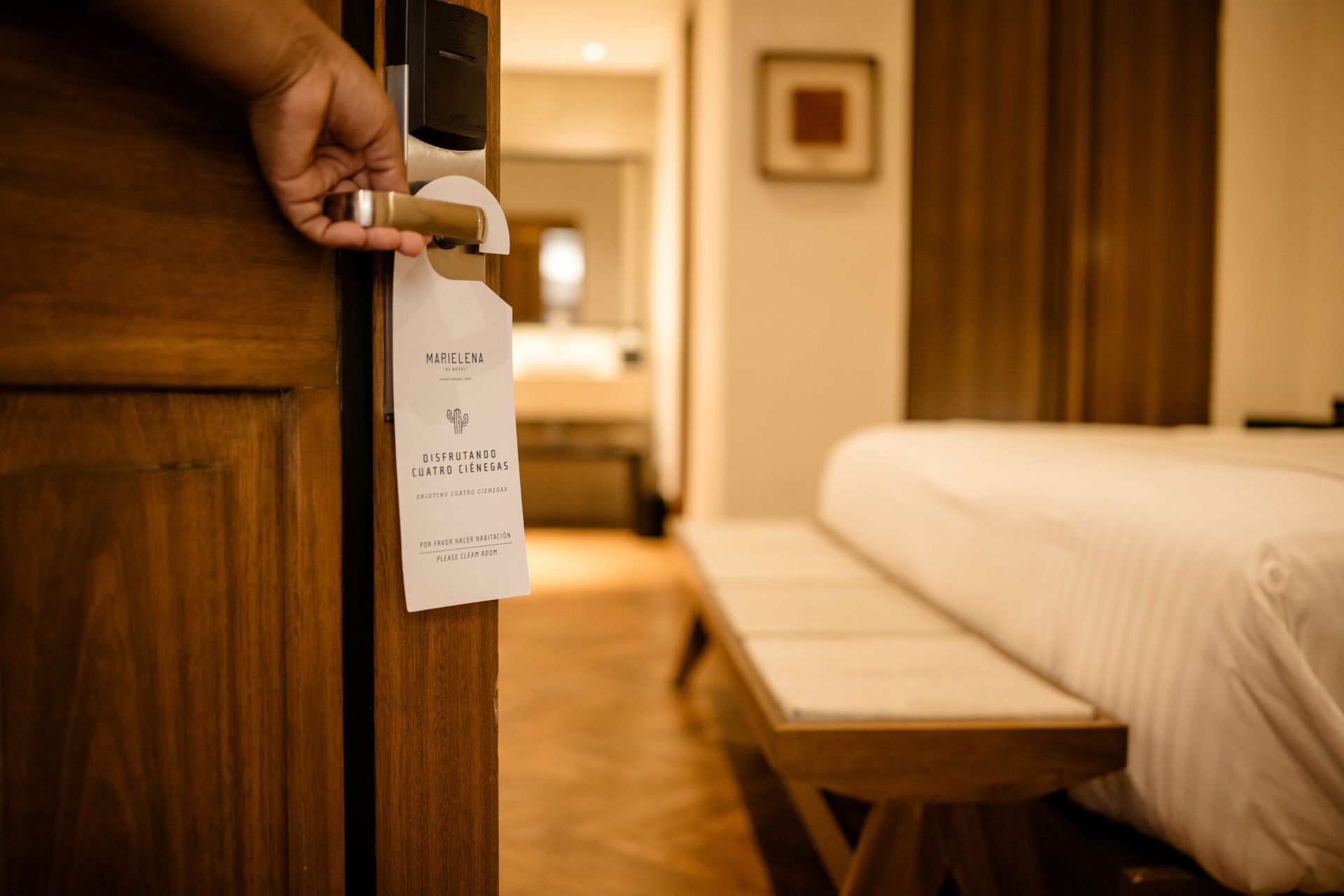 Una mano sostiene una etiqueta de puerta de hotel con una acogedora habitación y una cama impecablemente hecha al fondo.