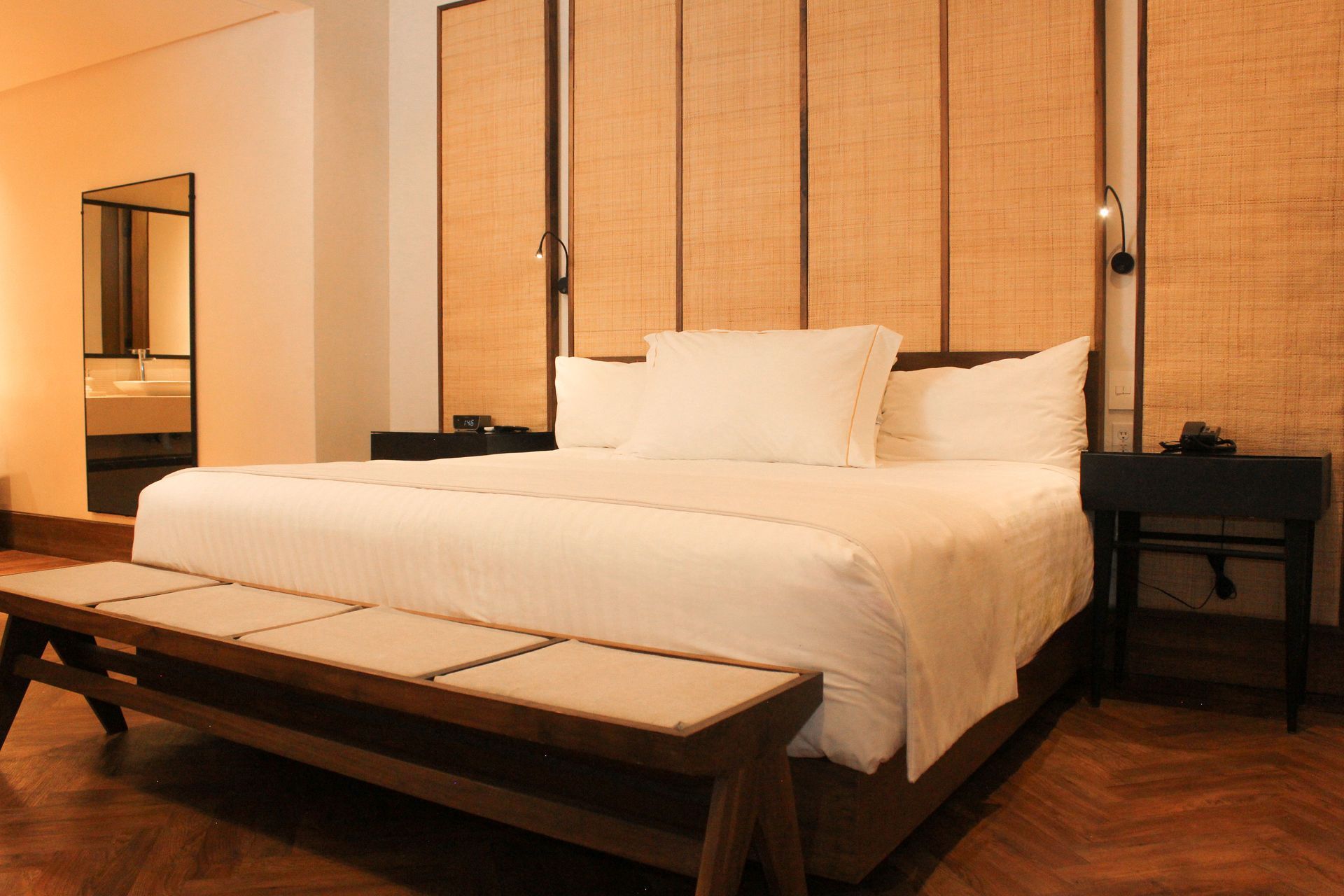 Dormitorio minimalista con cama blanca, banco de madera y paneles de pared tejidos.