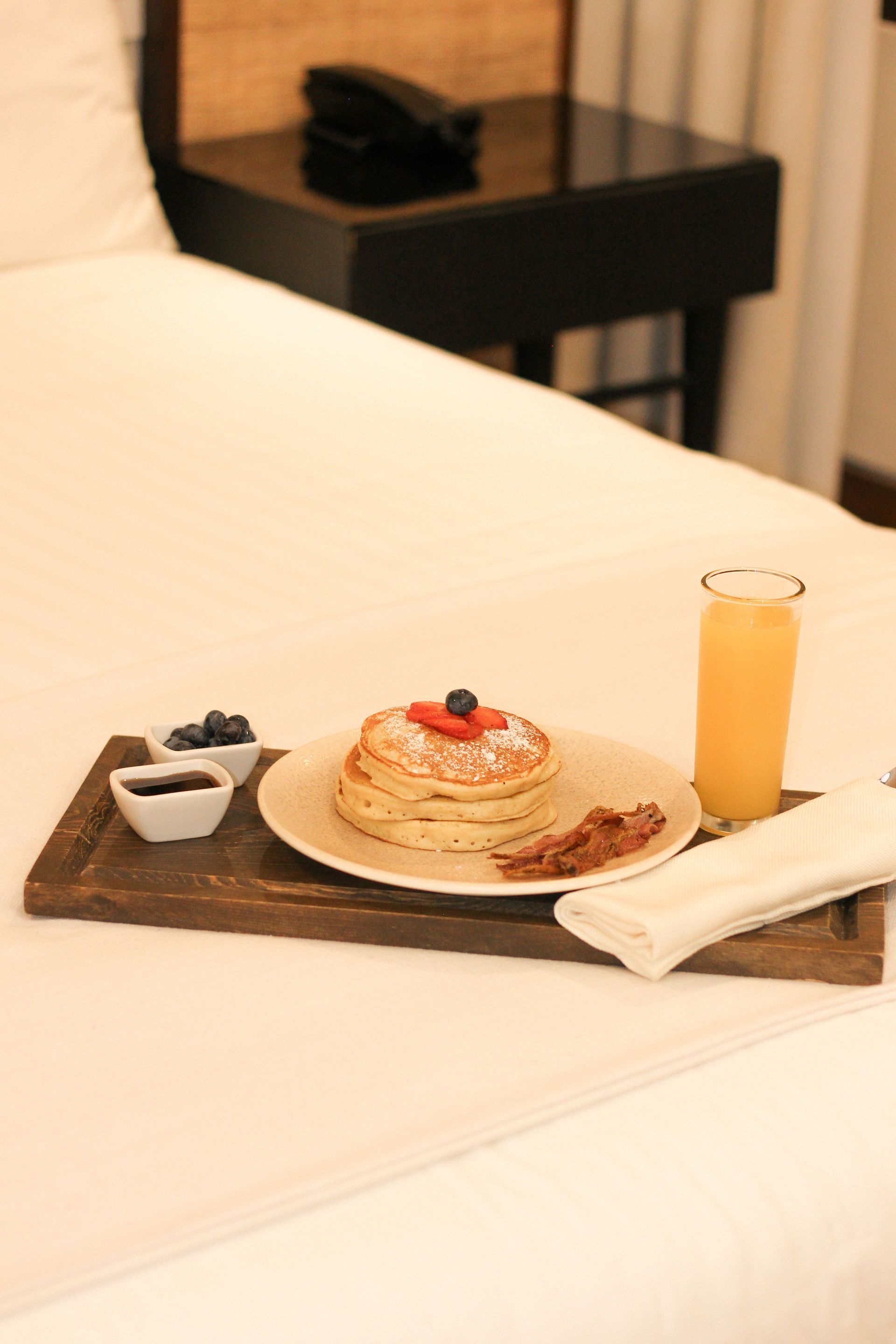 Bandeja de desayuno con panqueques, bayas, jugo de naranja y una servilleta sobre la cama.
