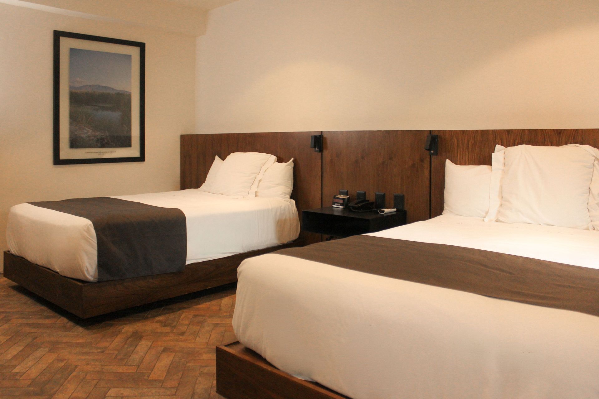 Habitación de hotel con dos camas bien hechas, una mesita de noche y un cuadro enmarcado en la pared.