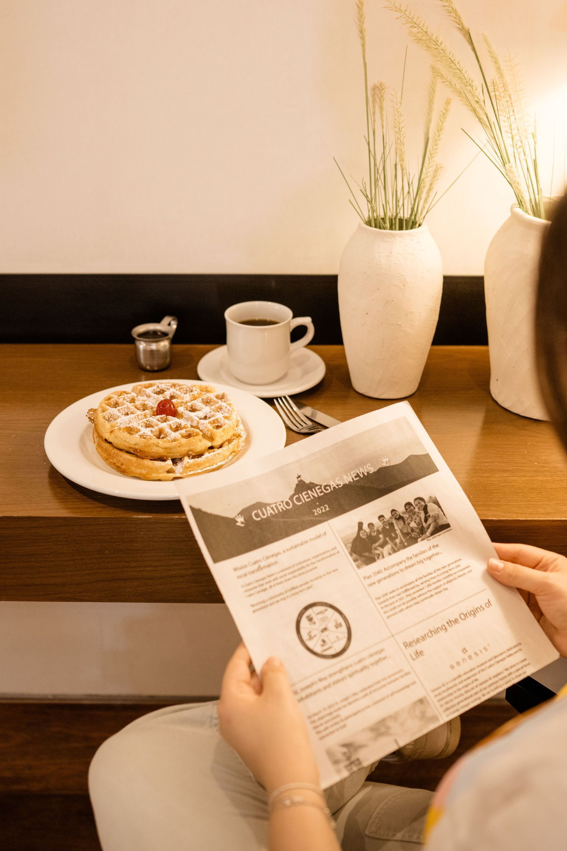 Una persona lee el periódico en una mesa de café con café, pasteles y jarrones blancos.