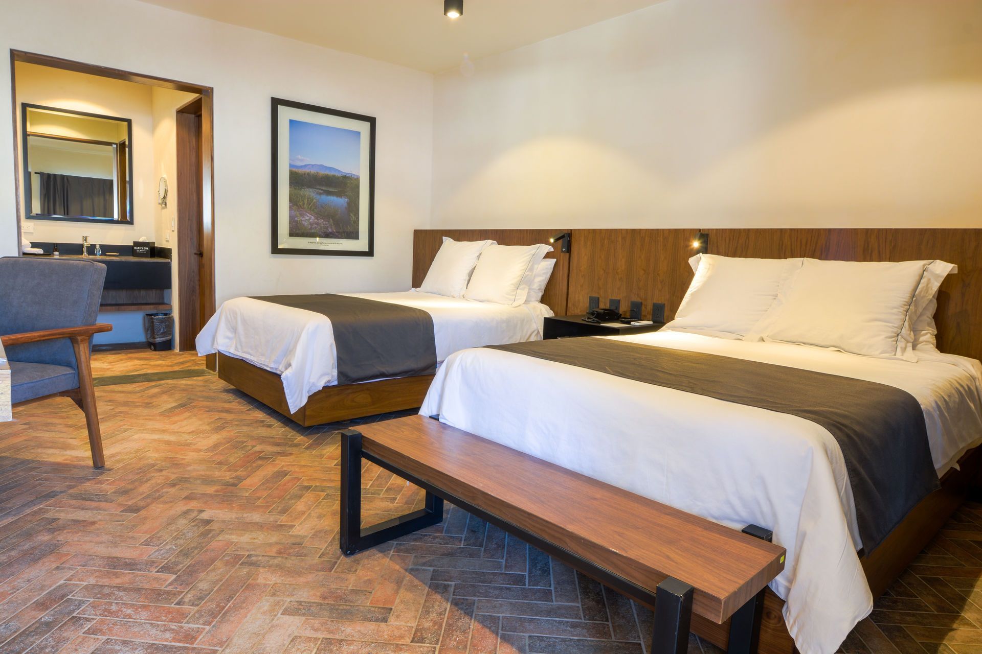 Habitación de hotel con dos camas bien hechas, un banco de madera y una silla de escritorio cerca de un espejo de pared.