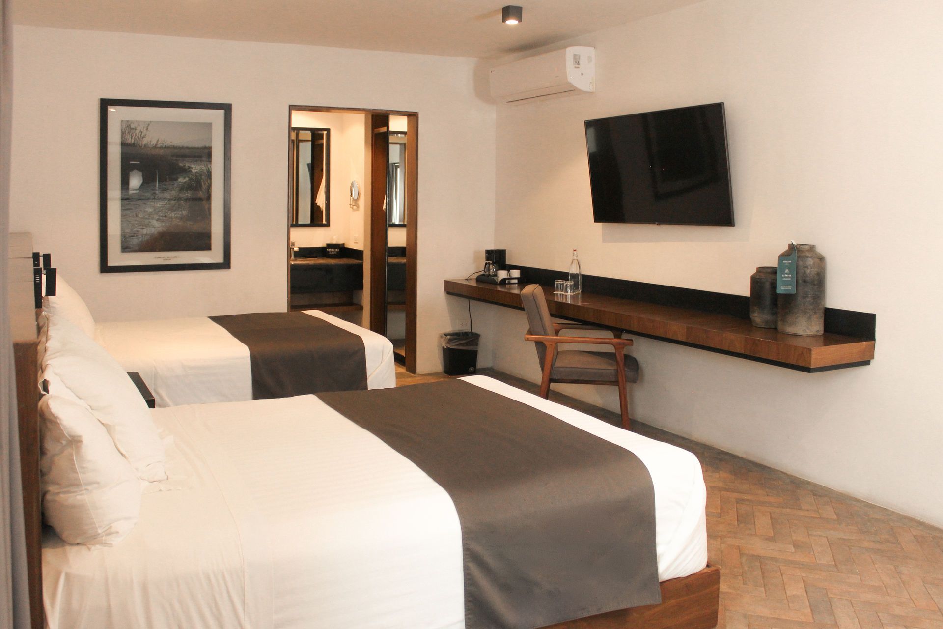 Habitación de hotel con dos camas, televisor montado en la pared, escritorio y puerta abierta al baño.