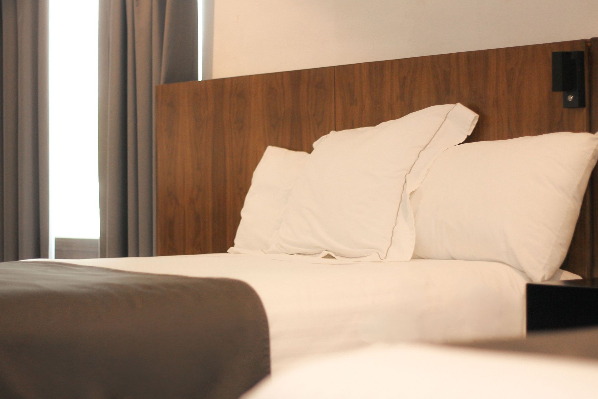 Cama de hotel bien hecha con almohadas blancas, manta gris y cabecero de madera junto a una ventana con cortinas.