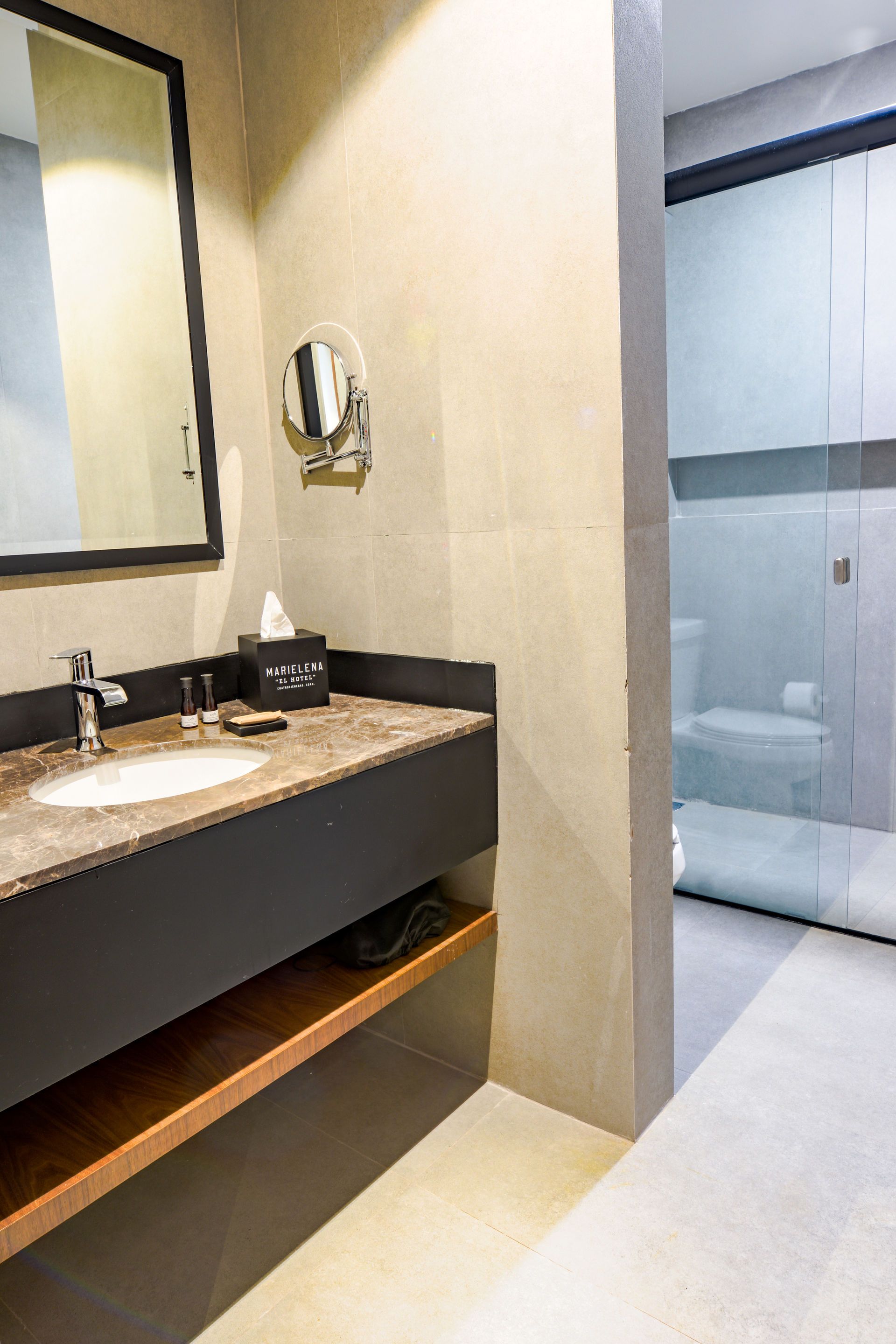 Tocador de baño moderno con lavabo, espejo y puerta de ducha de cristal sobre un suelo de baldosas claras.