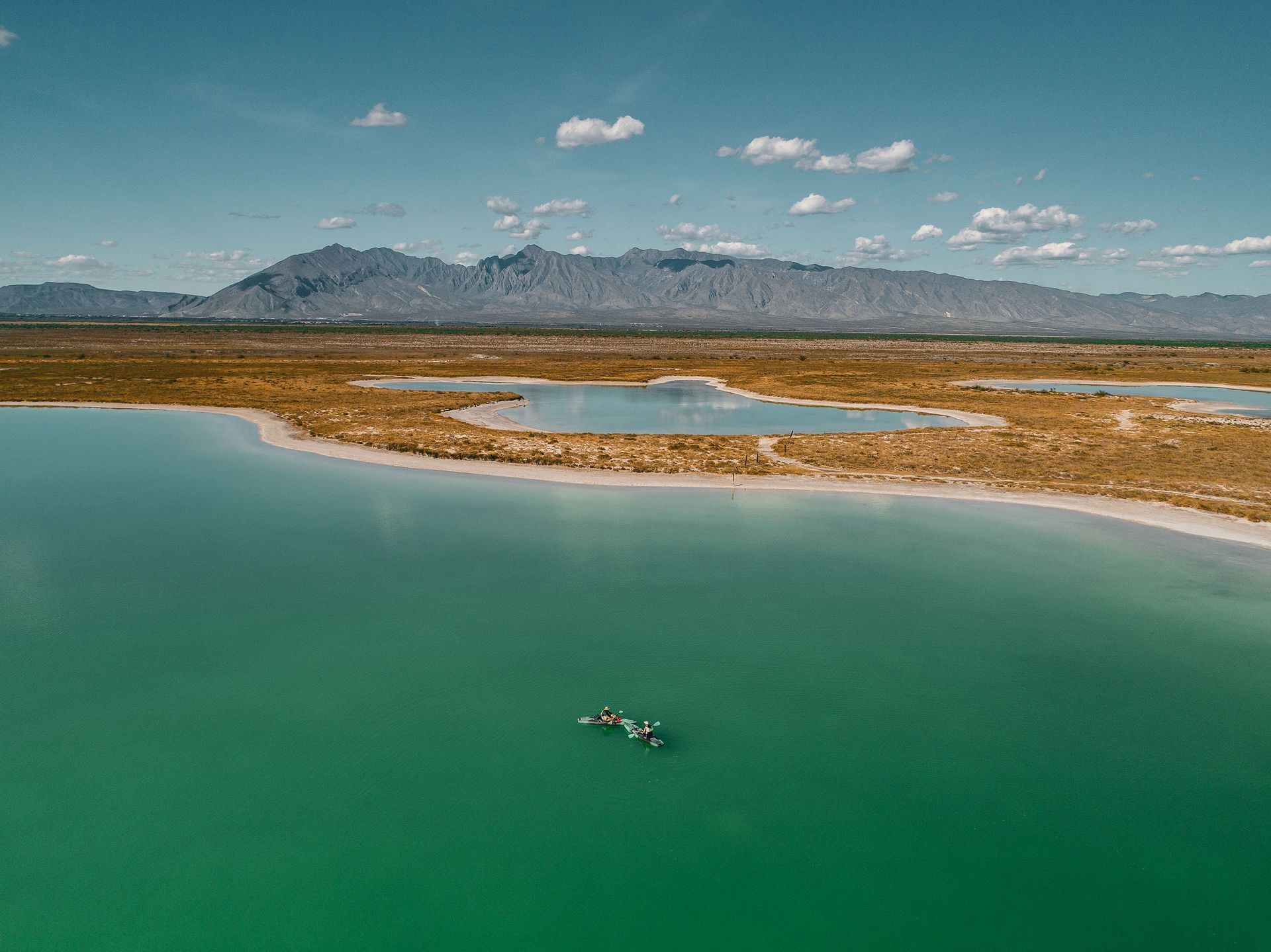 Lago azul verdoso con hierbas de marisma frente a una cordillera brumosa bajo un cielo despejado.