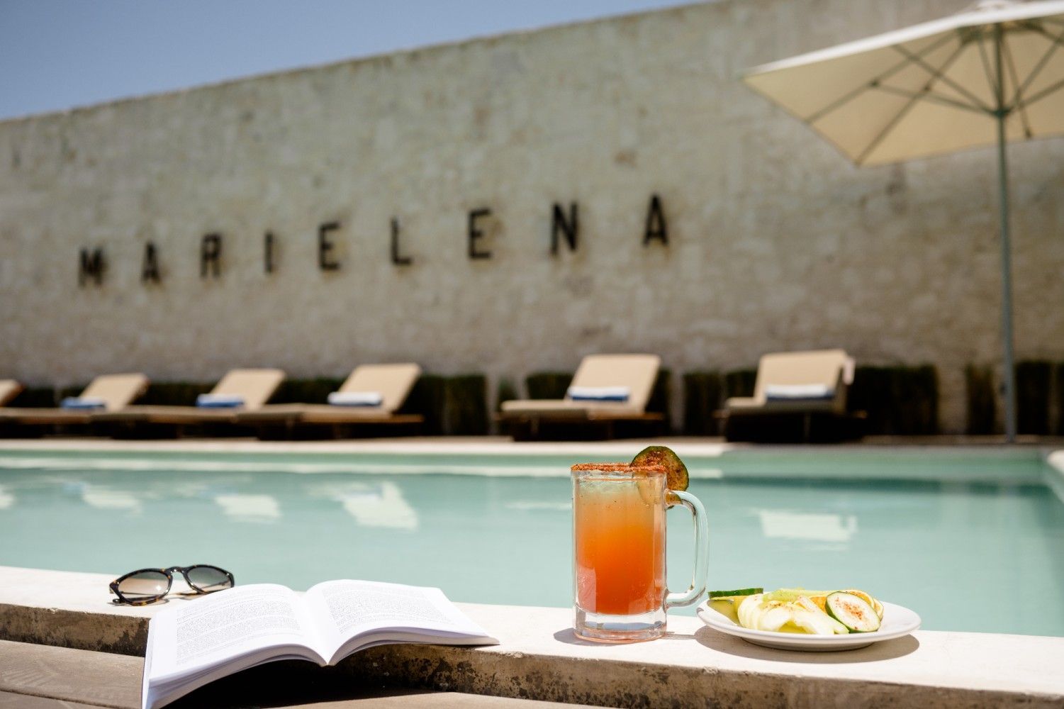 Desayuno junto a la piscina con bebida helada, libro abierto y gafas de sol en una soleada piscina de hotel.