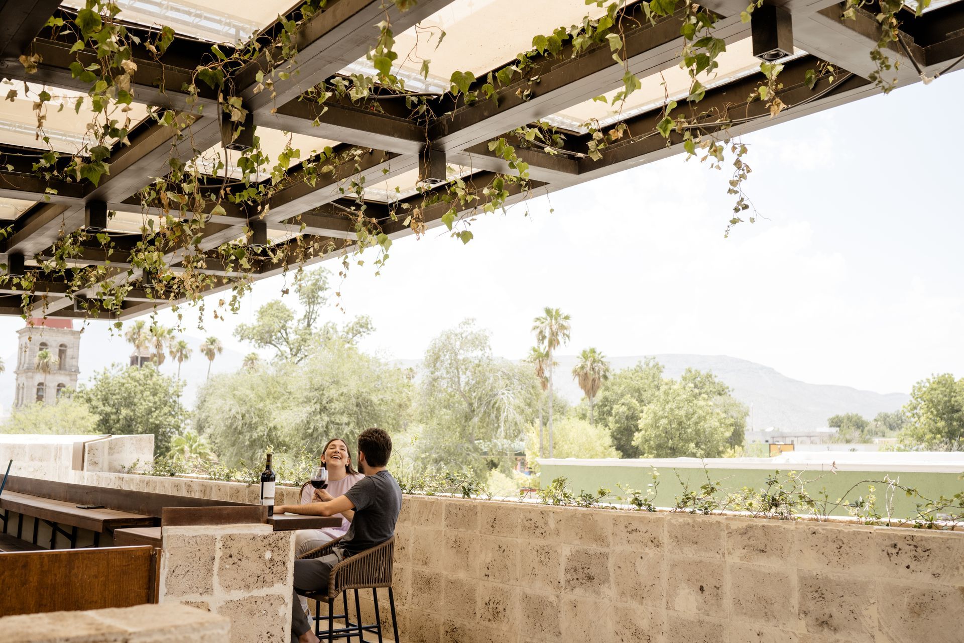 Persona sentada en una mesa de patio bajo una pérgola cubierta de enredaderas, con vistas a un luminoso jardín.