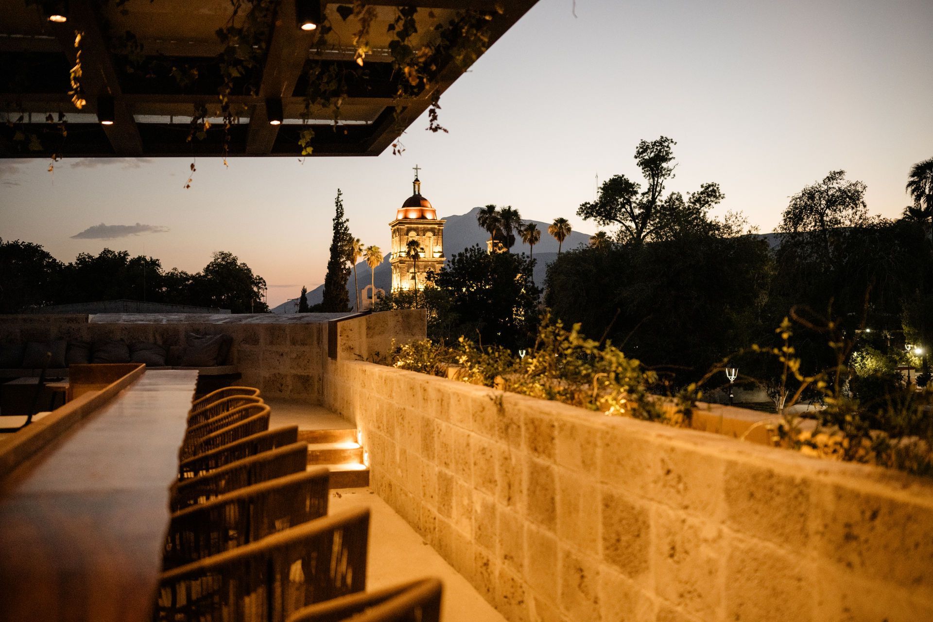 Terraza al atardecer con vistas a la cúpula iluminada de una iglesia y a los árboles que se extienden más allá de un muro.
