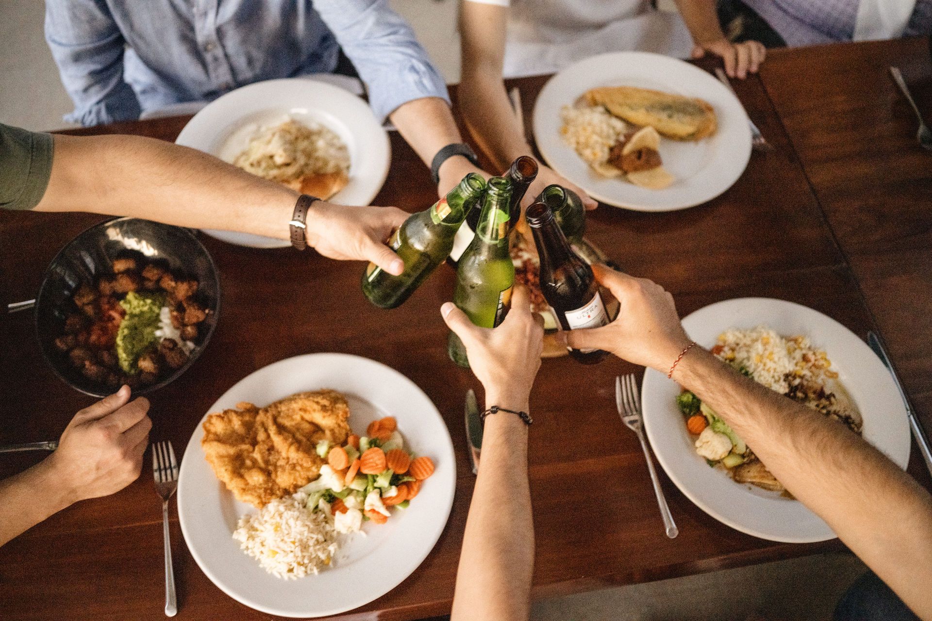 Un grupo de amigos choca sus botellas de cerveza alrededor de una mesa con platos de pasta y comida en un ambiente informal.