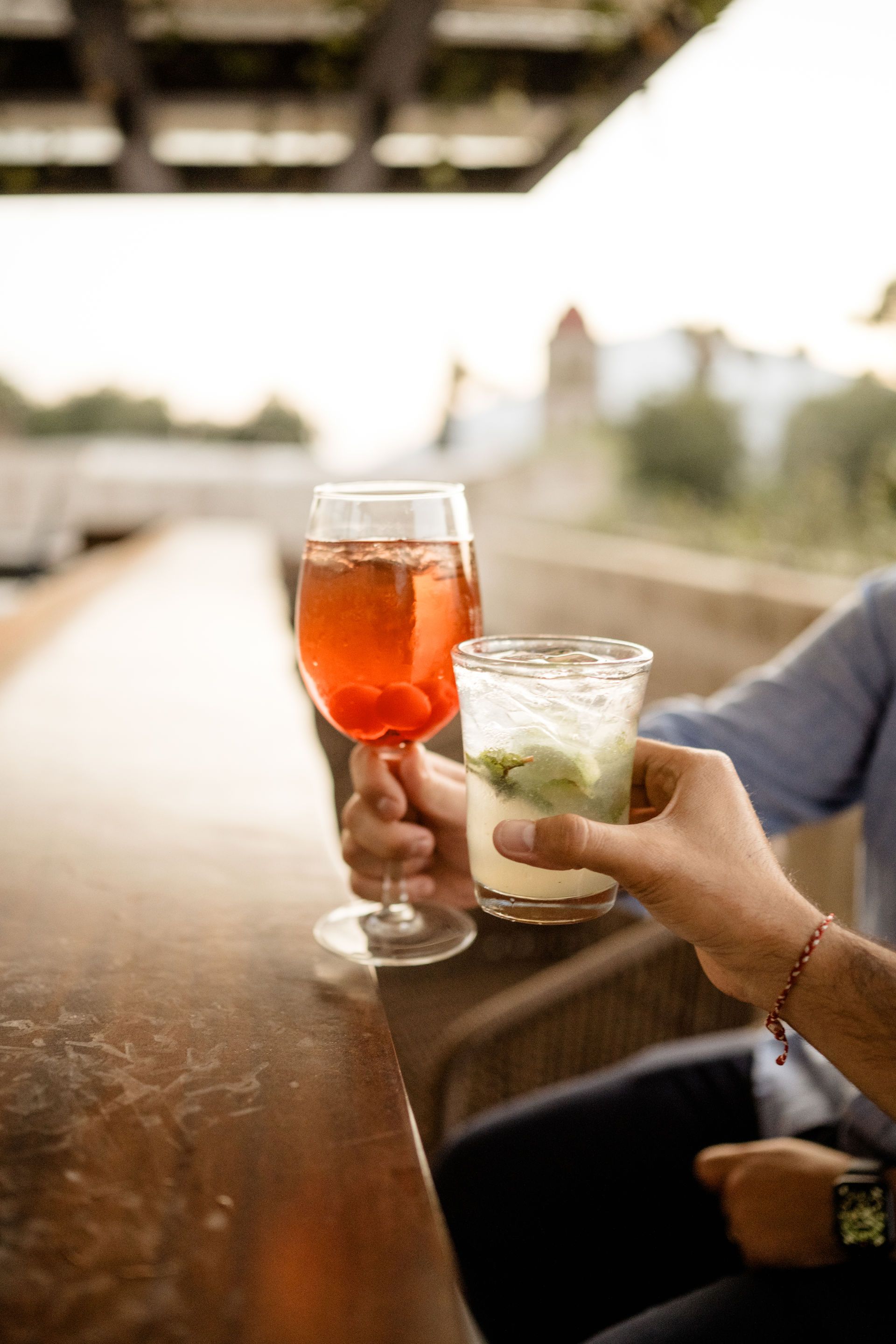 Manos que chocan con sus bebidas en una mesa de madera, con un cóctel rojo y una bebida de menta al aire libre.