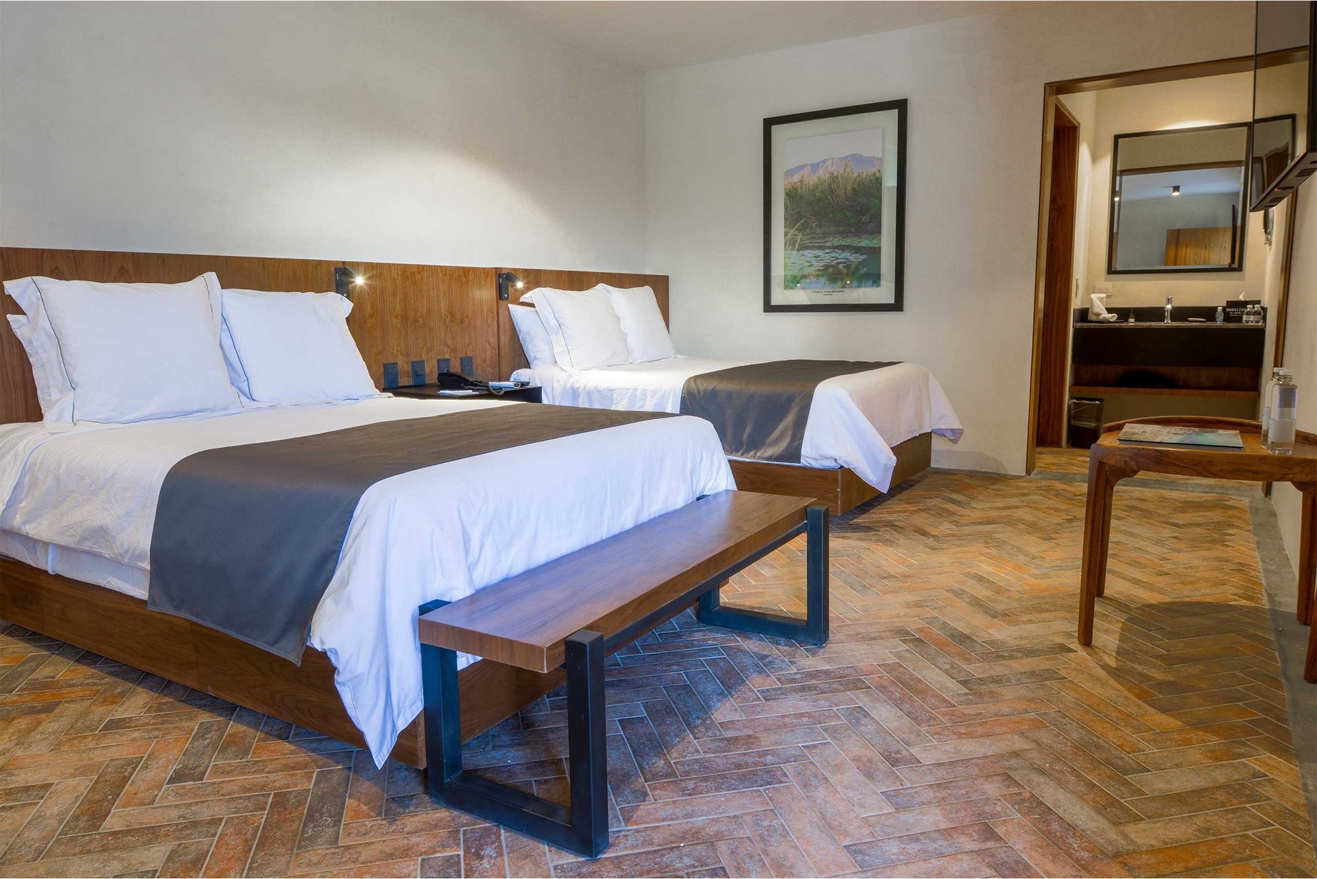 Habitación de hotel con dos camas hechas, banco de madera y decoración neutra.